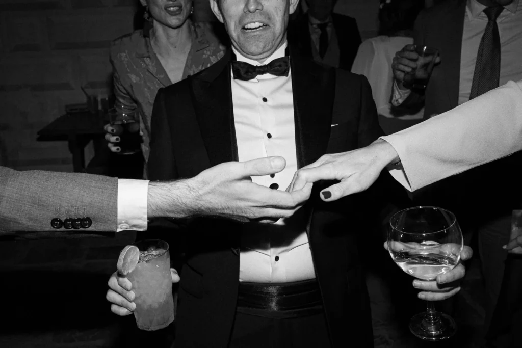 Guest in a tuxedo laughing as two hands meet in front of him at a wedding at Casa de Pilatos, Seville, black and white